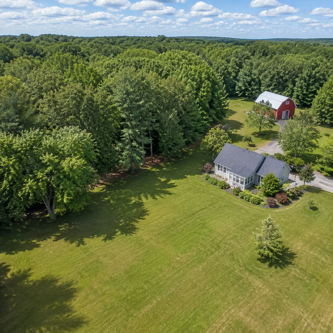 Aerial view of a property with open lawn, wooded area, house, and barn under partly cloudy sky.